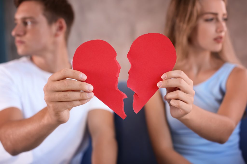 Young couple holding halves of broken heart on sofa at home. Relationship problems