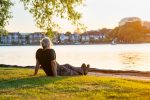Back view gray hair mature man sitting on the green grass at the bank of park's lake and enjoying the sunset.