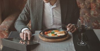 man uses his tablet while eating healthy breakfast shakshuka - fried eggs, tomatoes and spices in cast iron stewpan on old wooden plank in an indoor restaurant.