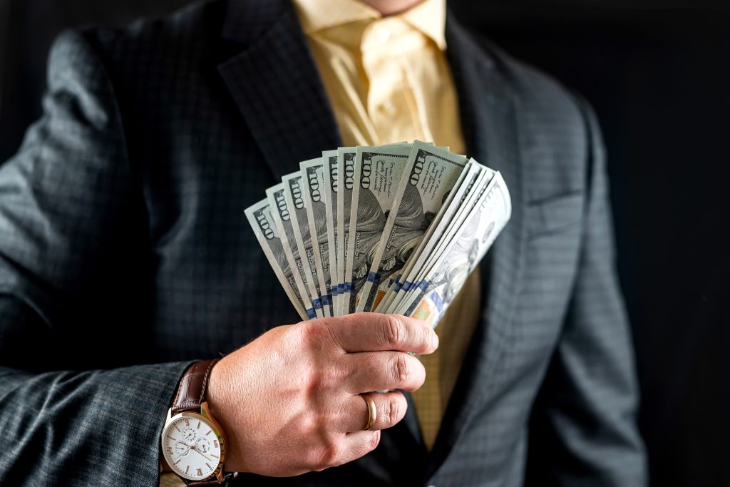 businessman in a shirt and jacket counts hundred dollar bills in front of his chest.
