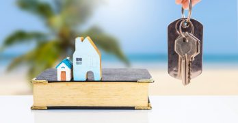 Miniature house on old book with house key in girl hand over blurred tropical beach background, property and real estate investment business, house insurance